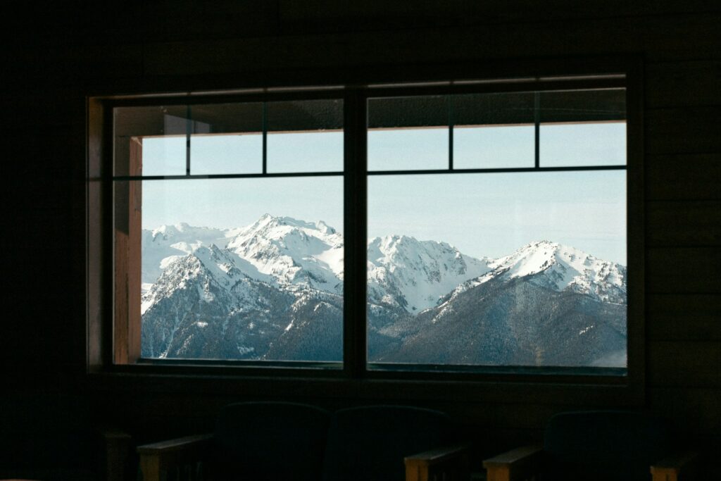 View Of Snow-Covered Mountains Framed By A Large Window, With Dark Furniture Silhouettes In The Foreground. The Scene Evokes A Sense Of Tranquility And Highlights The Importance Of Knowing How To Replace Home Windows For Maintaining Such Picturesque Views.